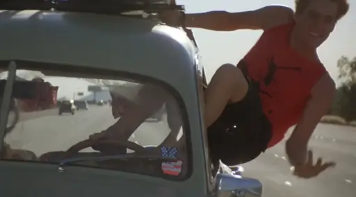 Jim Carrey hanging out of the window of a moving vehicle, his feet controlling the steering wheel