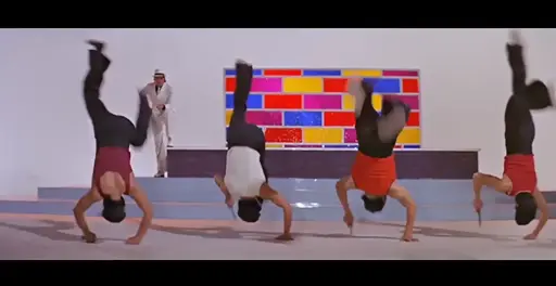 Four men doing handstands, some with a knife in one hand, in front of a coloured background & a man in a white suit & hat